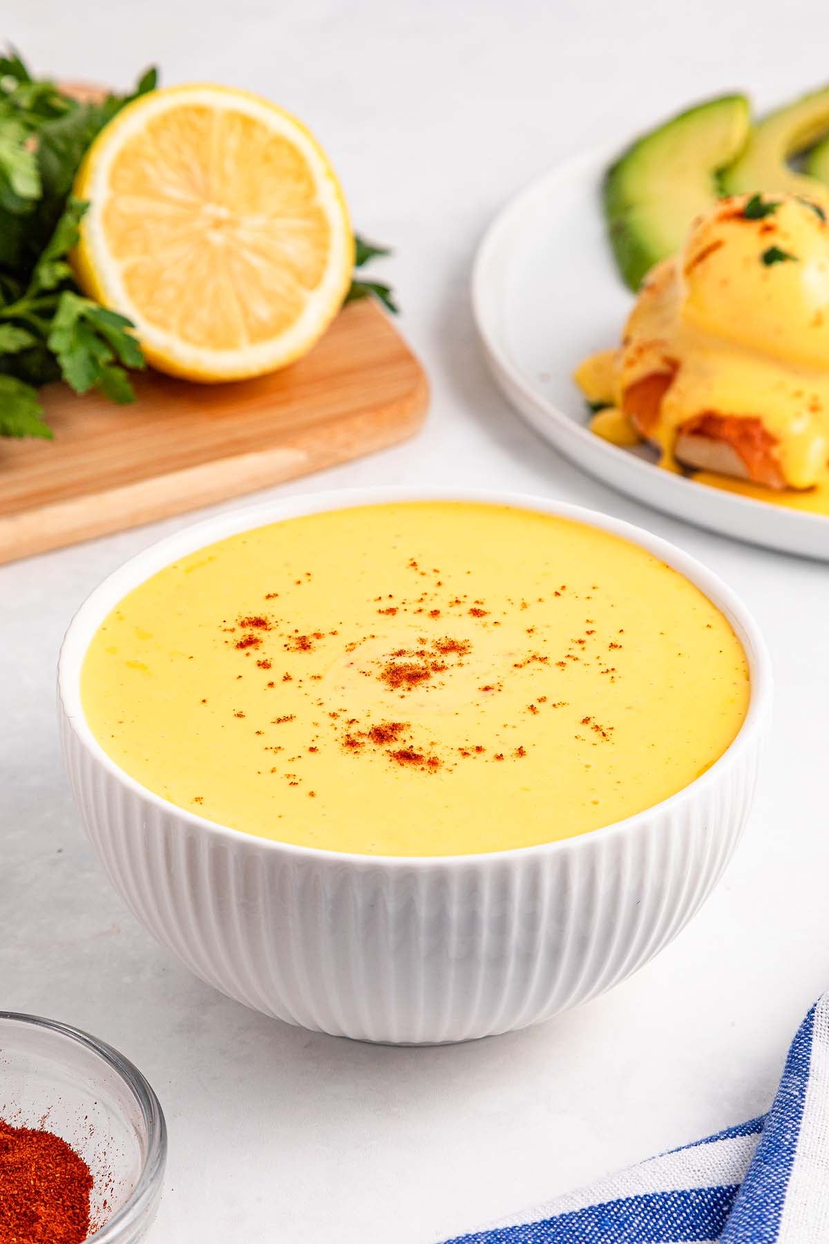 A bowl of creamy yellow Easy Hollandaise Sauce, sprinkled with paprika, sits on a white surface near fresh ingredients.