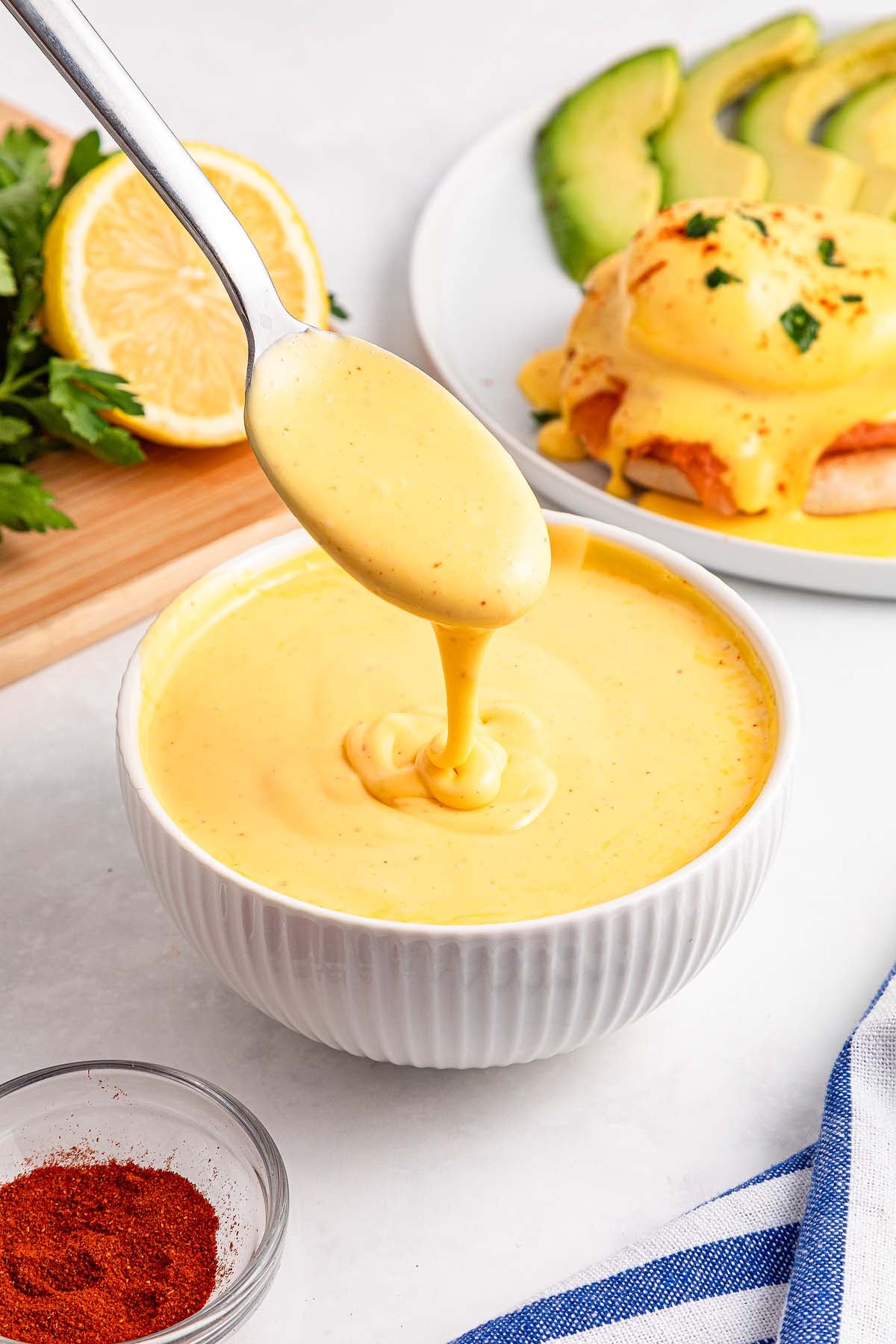 Spoon drizzling creamy yellow Easy Hollandaise Sauce into a white bowl, with eggs Benedict in the background.