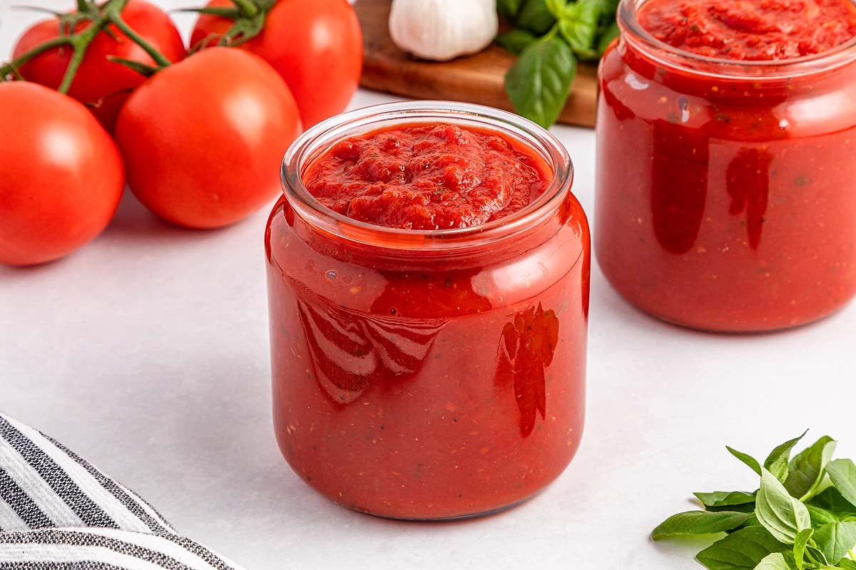 Glass jars filled with easy marinara sauce, surrounded by fresh tomatoes, basil, and garlic on a white surface.