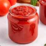 A glass jar filled with easy marinara sauce sits on a white surface, with fresh tomatoes and basil nearby.