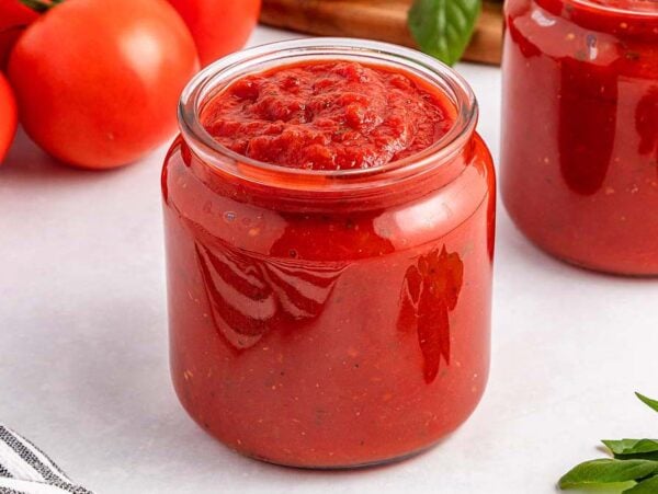 A glass jar filled with easy marinara sauce sits on a white surface, with fresh tomatoes and basil nearby.