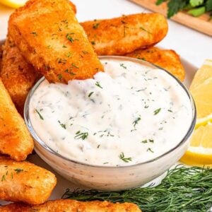 Golden fried fish sticks with dill served with easy tartar sauce and lemon wedges on a white plate.