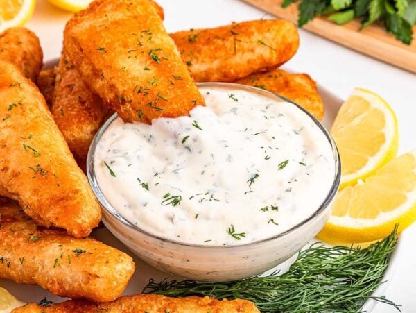 Golden fried fish sticks with dill served with easy tartar sauce and lemon wedges on a white plate.