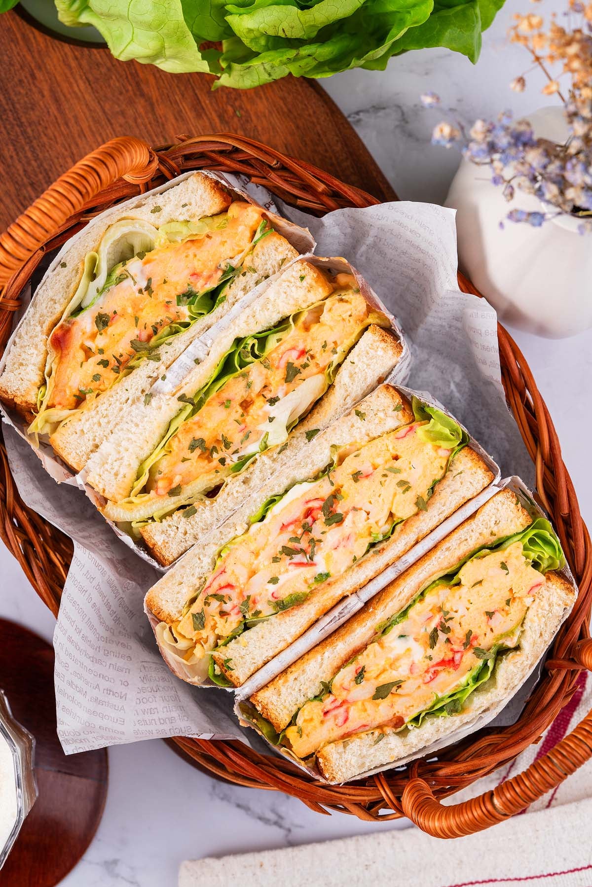 Four thick crab sandwiches with lettuce and egg salad filling, arranged in a wicker basket lined with paper.