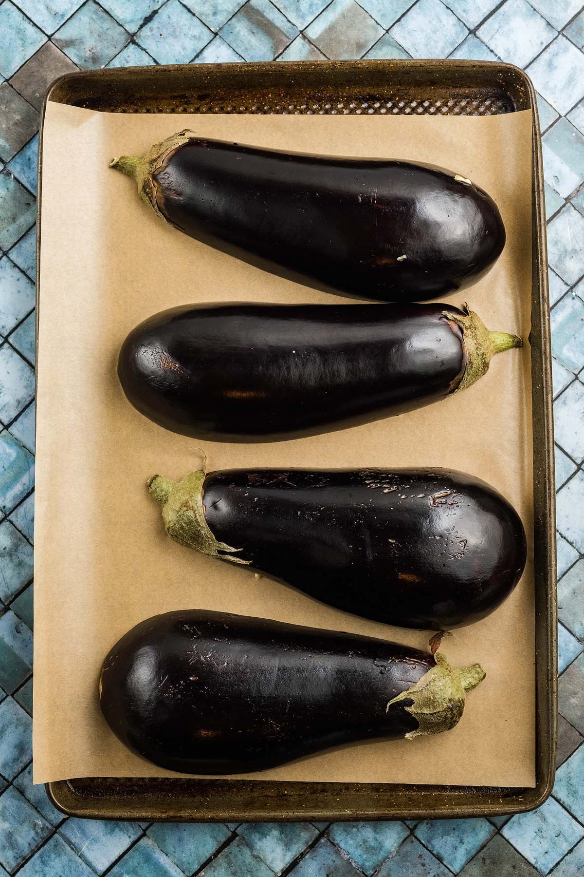 Four whole eggplants, ideal for making stuffed eggplant, rest on a parchment-lined baking sheet over a blue tiled surface.