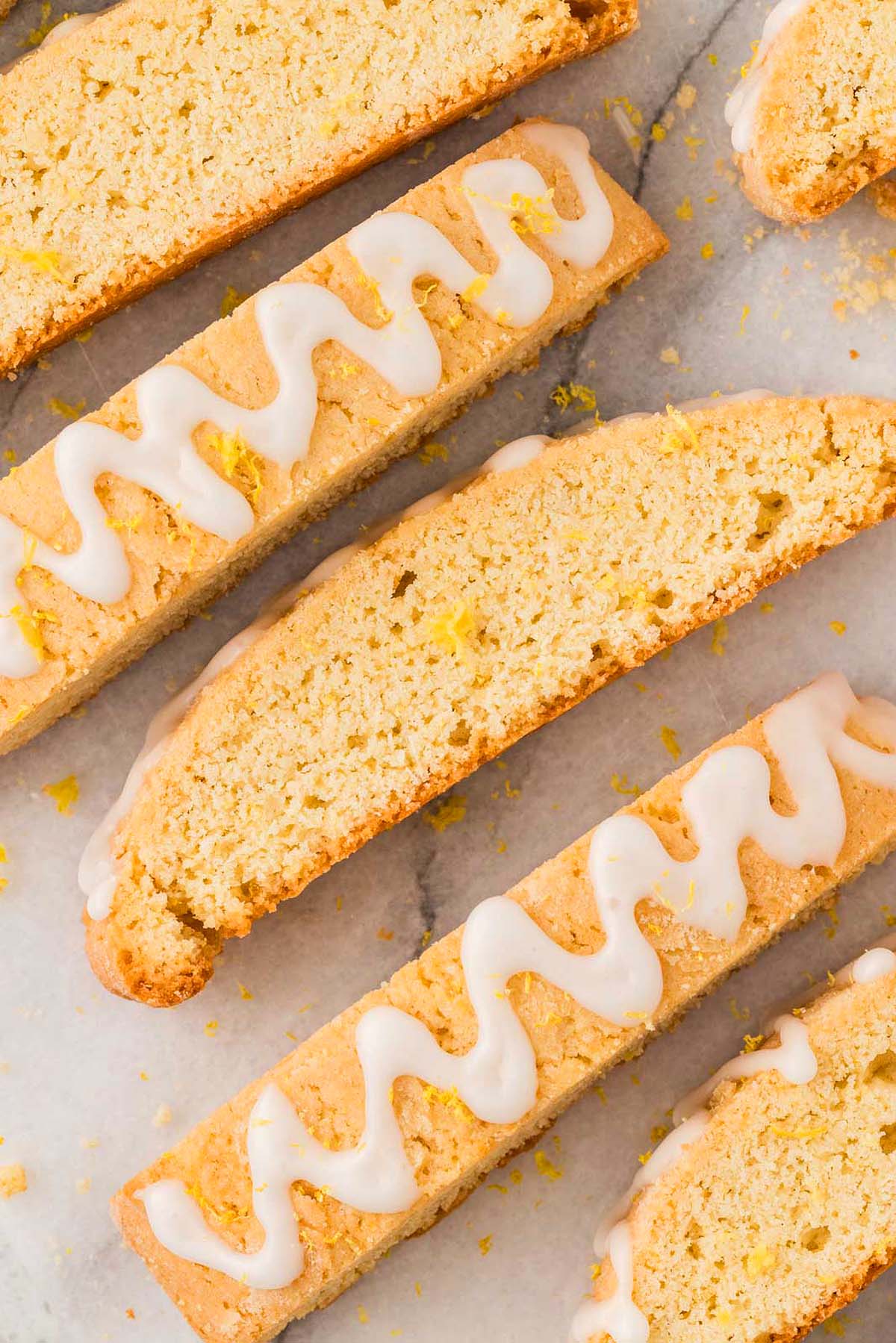 Four lemon biscotti, some drizzled with white icing, arranged on a marble surface and sprinkled with fresh lemon zest.