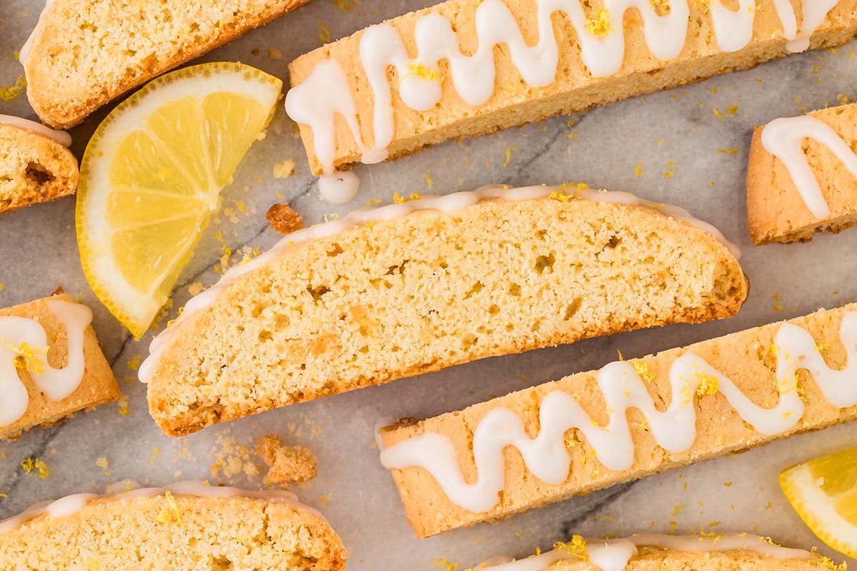 Lemon biscotti with a white icing drizzle and fresh lemon slices displayed on a marble surface.