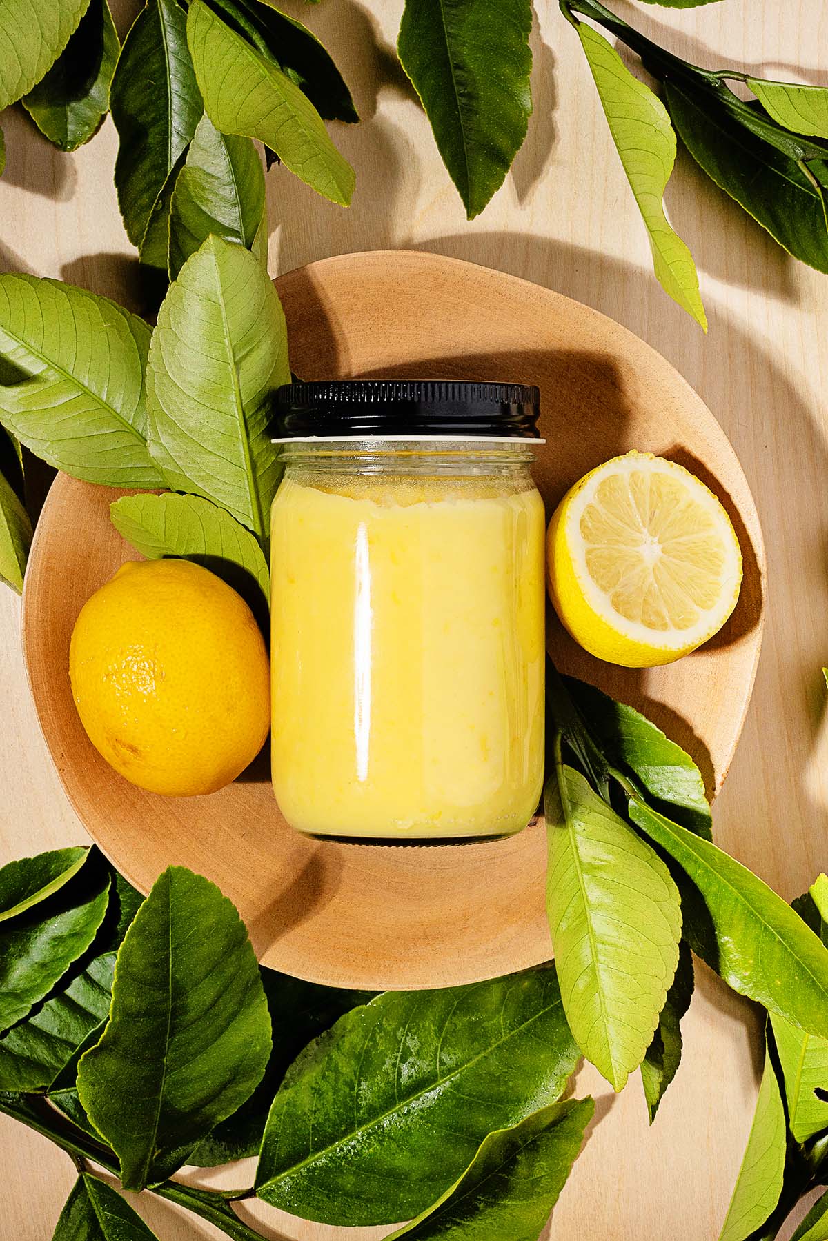 A jar of lemon curd sits in a wooden bowl, surrounded by a whole lemon, a lemon half, and fresh green leaves.