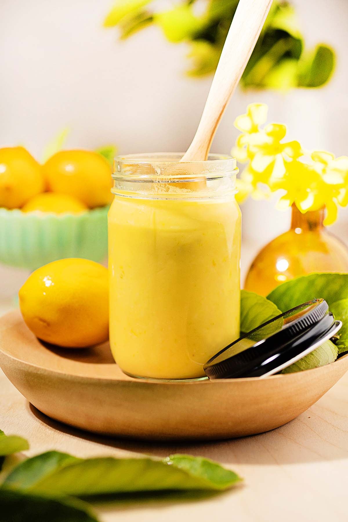 A jar of lemon curd with a wooden spoon, surrounded by fresh lemons and green leaves, captures the zesty essence of homemade lemon curd.