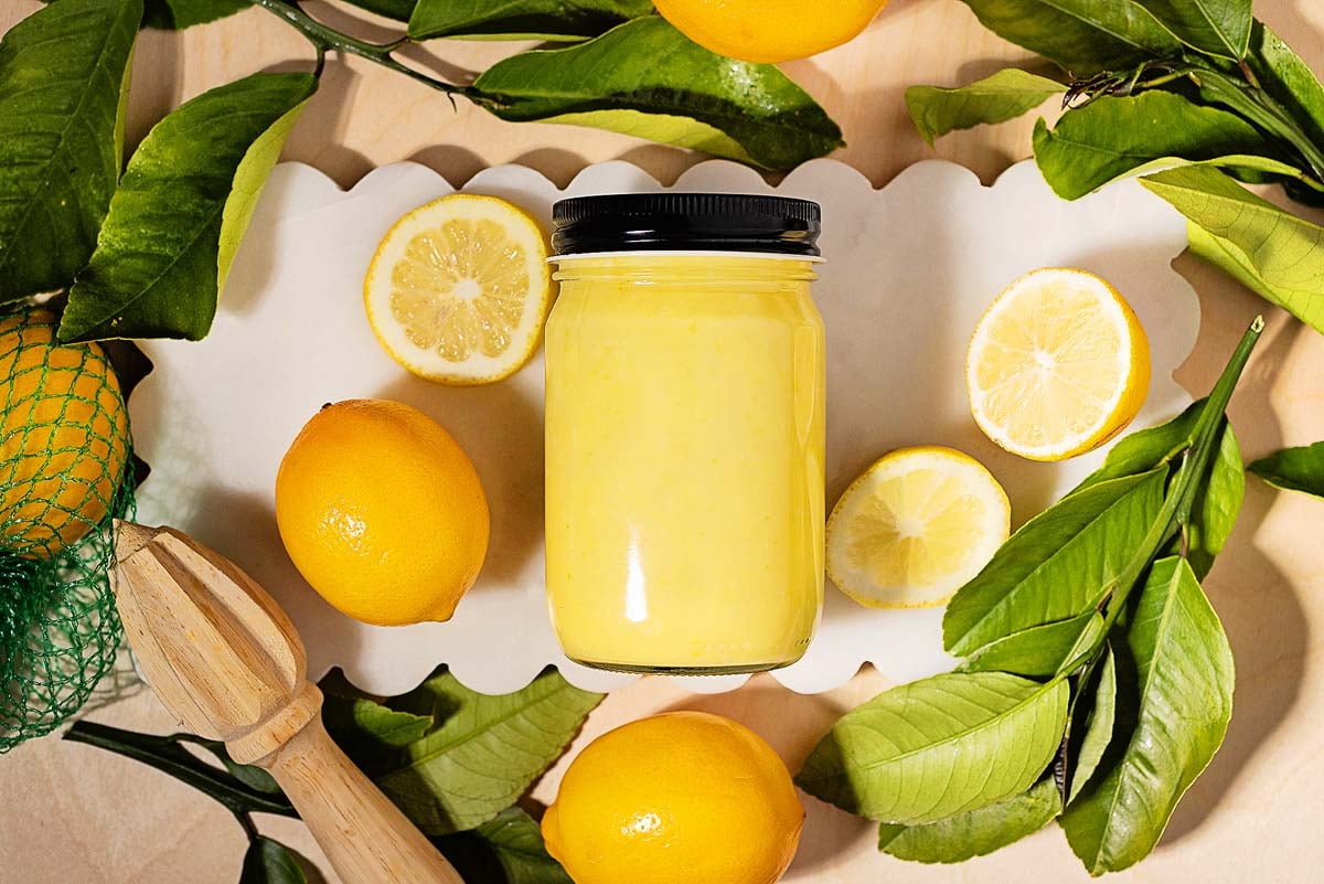 A jar of creamy lemon curd surrounded by fresh lemons, lemon leaves, and a wooden citrus reamer on a white tray.