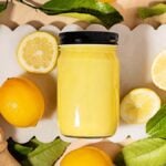 A jar of tangy lemon curd is surrounded by fresh lemons, lemon slices, and green leaves on a white scalloped tray.