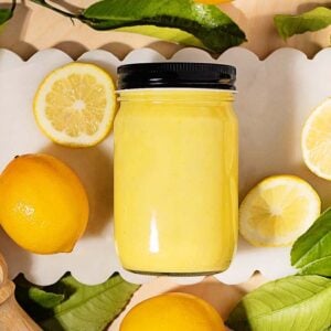 A jar of tangy lemon curd is surrounded by fresh lemons, lemon slices, and green leaves on a white scalloped tray.