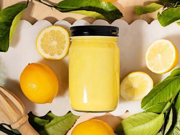 A jar of tangy lemon curd is surrounded by fresh lemons, lemon slices, and green leaves on a white scalloped tray.