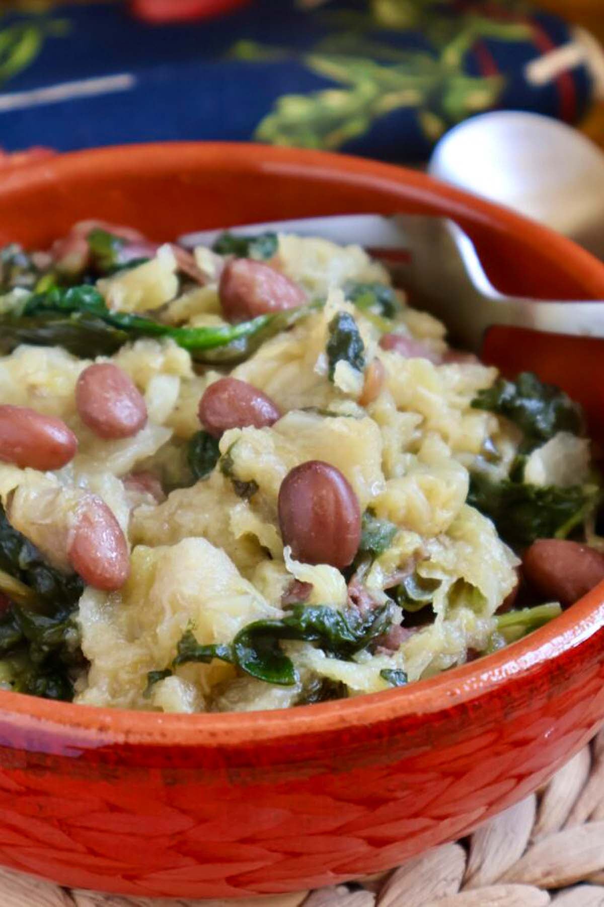 Mashed potatoes mixed with greens and red beans in a red bowl, reminiscent of hearty Italian soup recipes, with a spoon in the background.