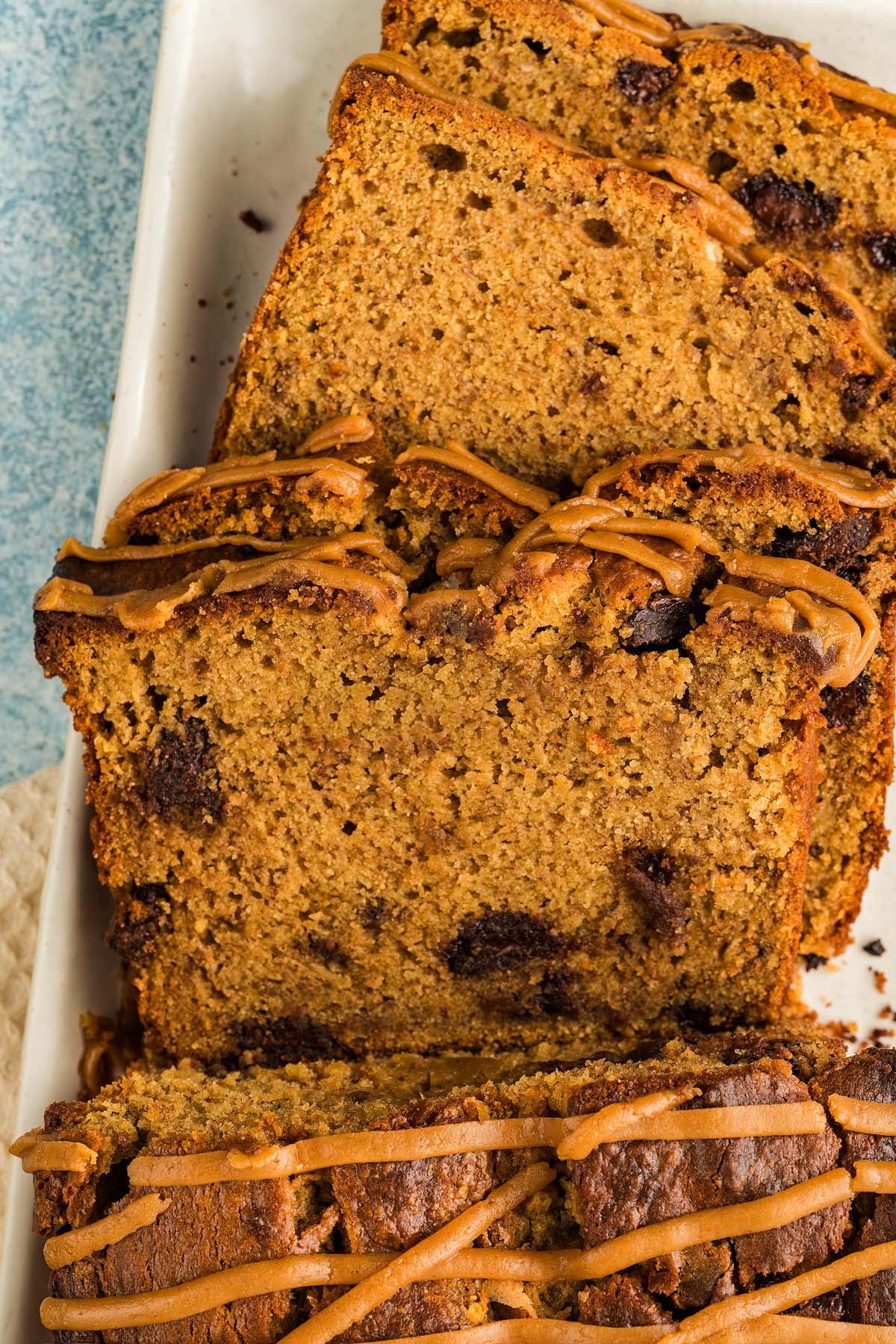 Sliced loaf of Peanut Butter Banana Bread with chocolate chips and a brown drizzle on top, arranged on a white plate.