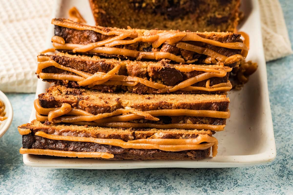 Three slices of chocolate chip Peanut Butter Banana Bread drizzled with caramel icing, stacked on a white rectangular plate.