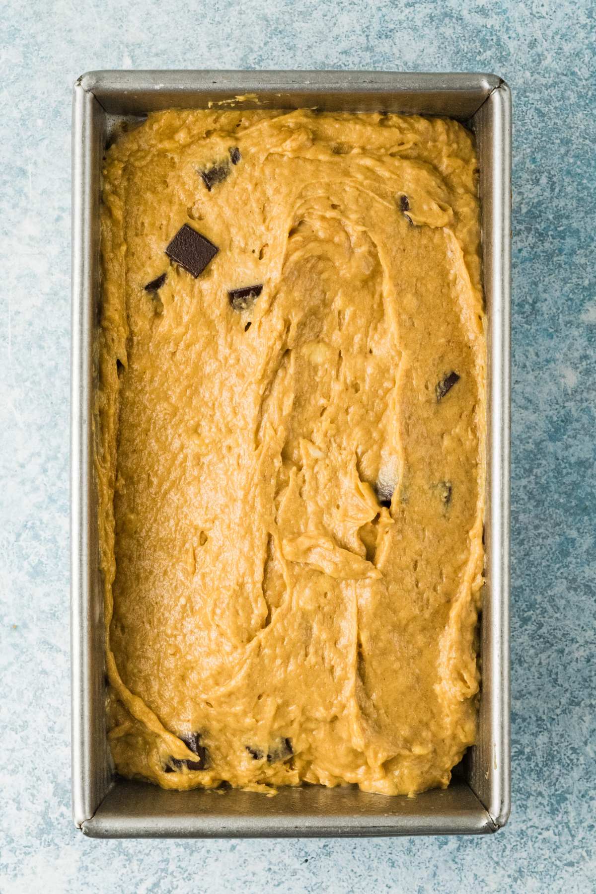 Banana bread batter with chocolate chunks in a loaf pan, ready to be baked, on a light blue surface.