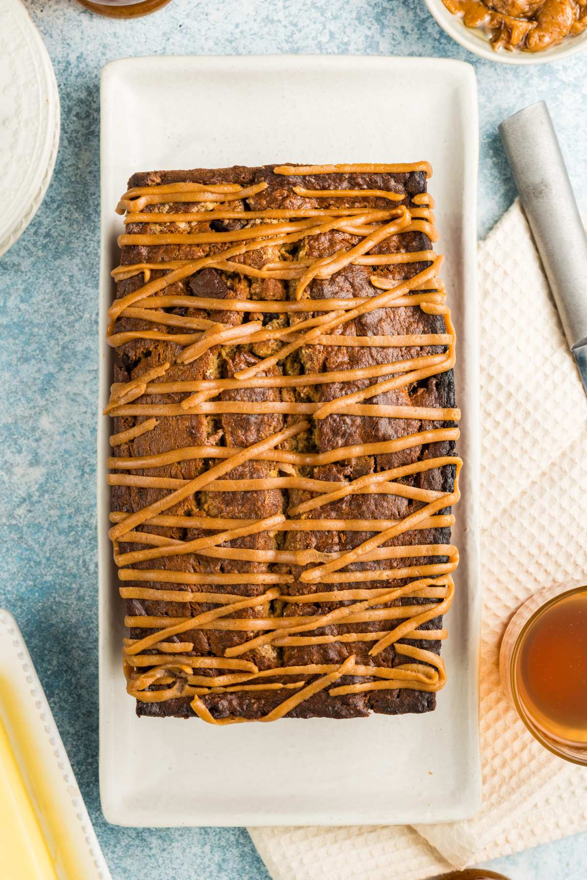 A loaf of bread drizzled with caramel sauce on a white rectangular plate, surrounded by kitchen items.