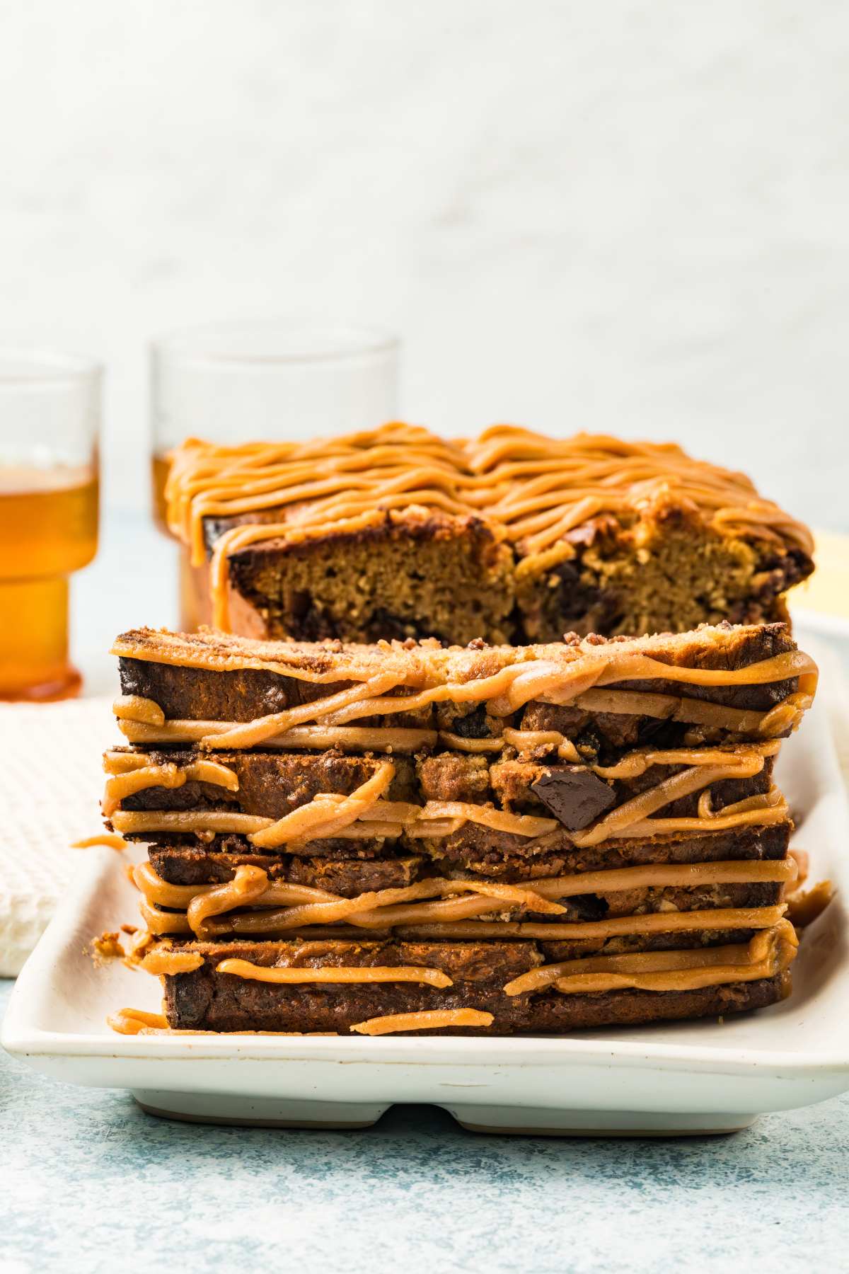 Sliced loaf of bread with caramel drizzle, stacked on a white plate, two drinks in the background.