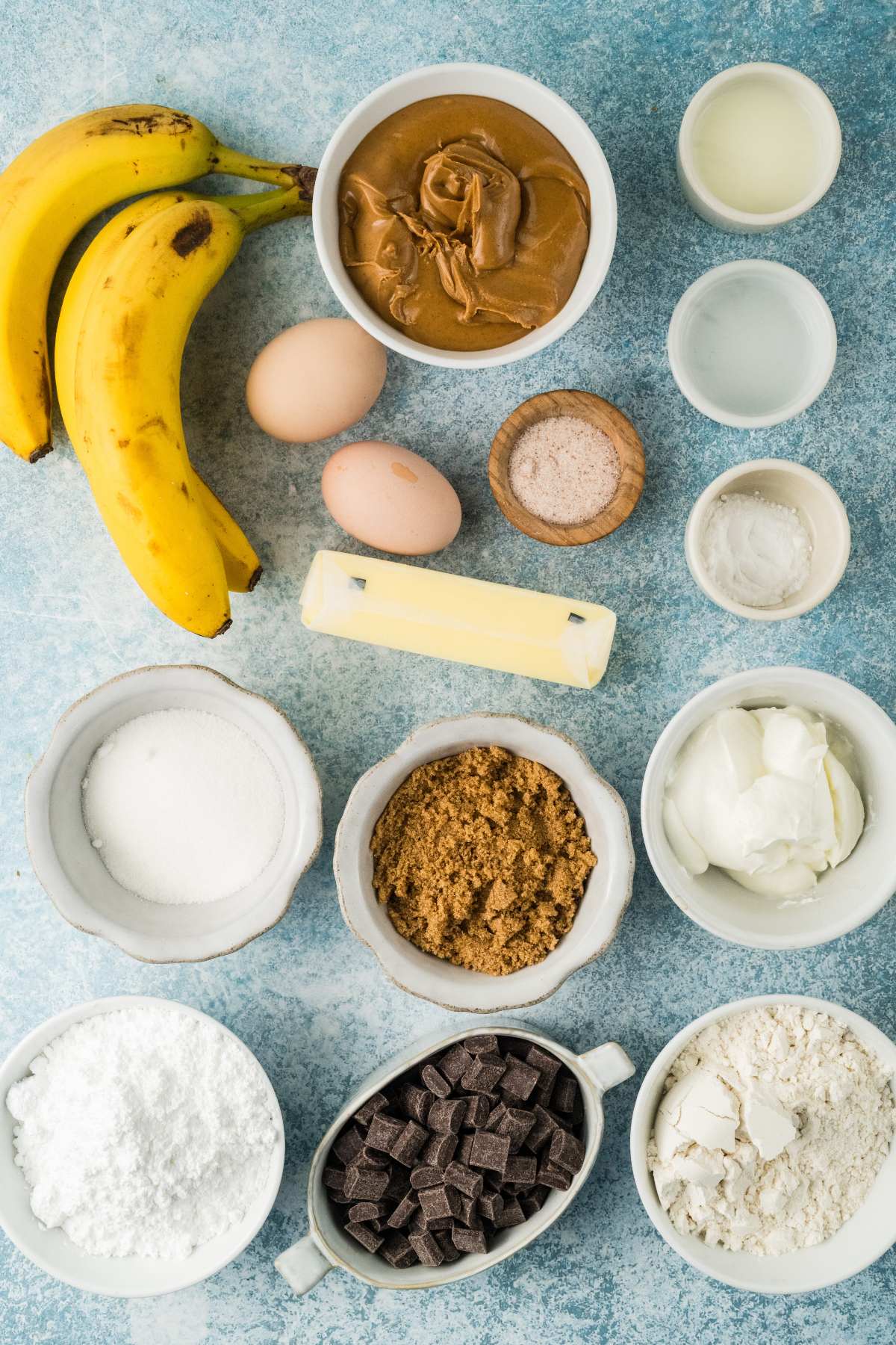 Bananas, eggs, peanut butter, sugars, flour, baking essentials, chocolate chunks, and butter on a blue surface.