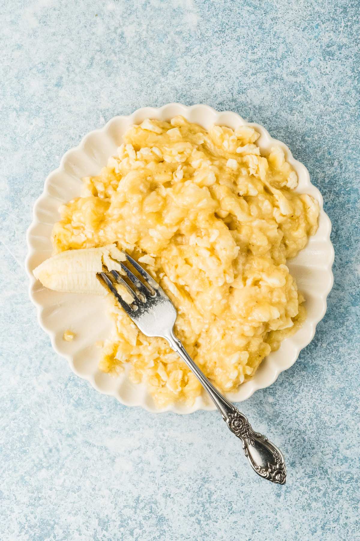 A white plate with mashed bananas and a fork on a light blue textured surface.