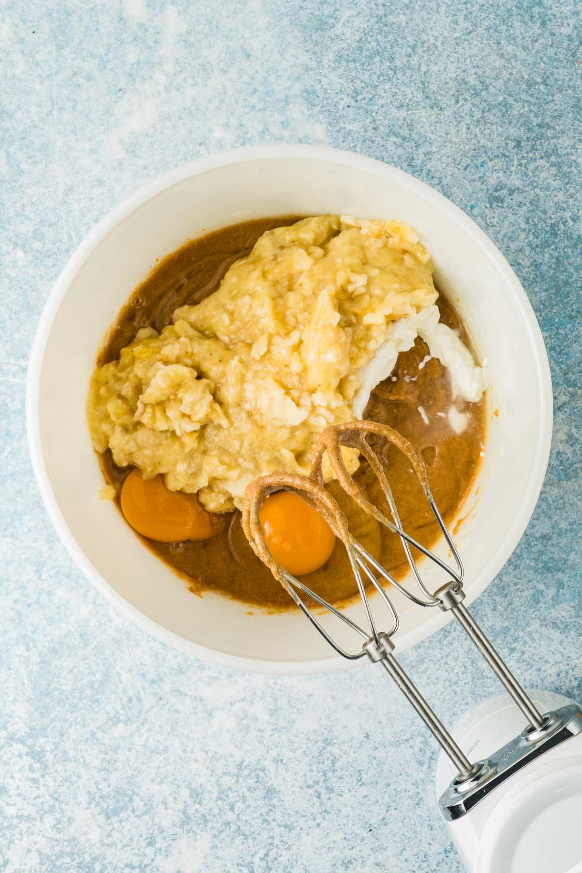 A mixing bowl with mashed banana, eggs, and batter, beside a hand mixer on a light blue surface.