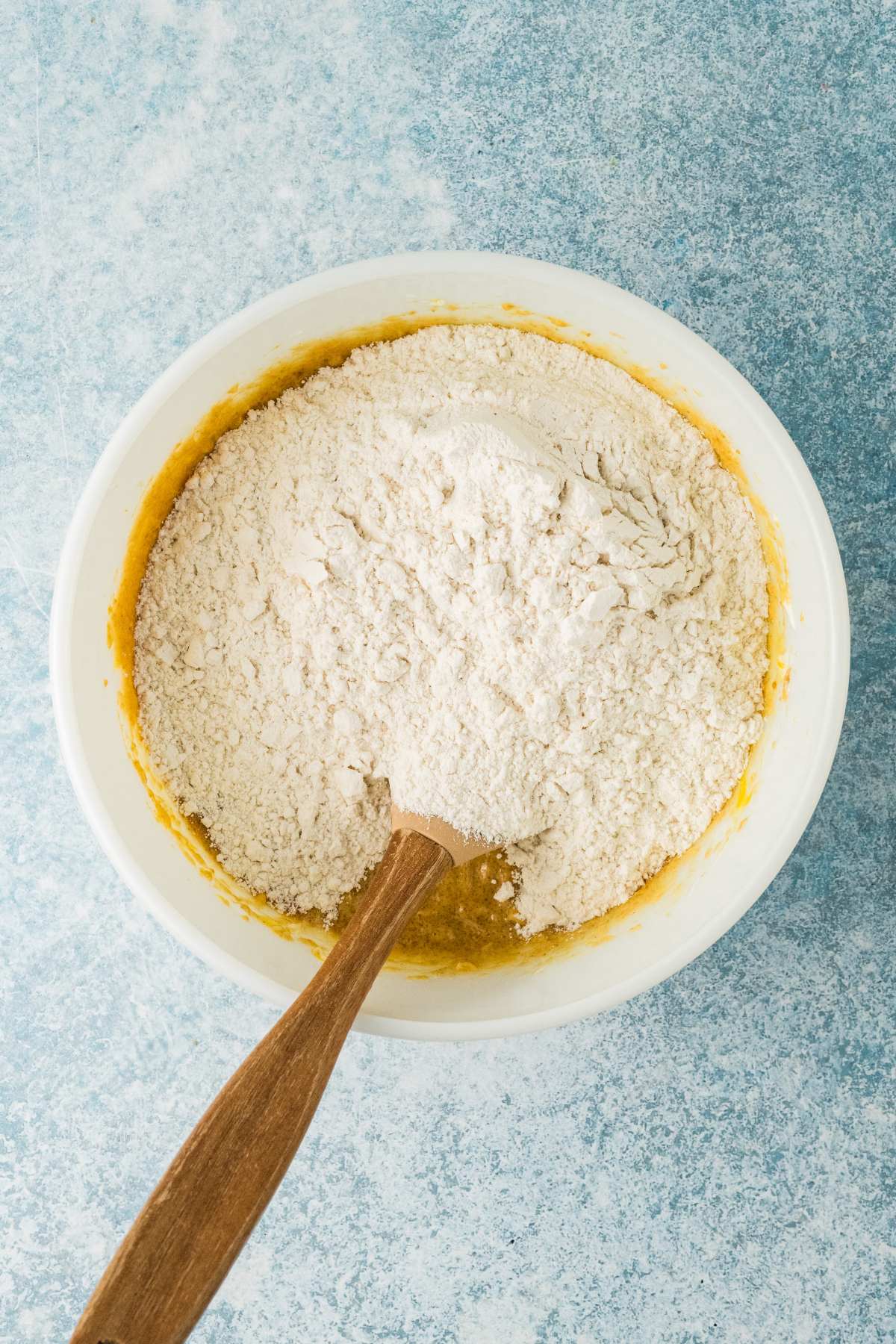 A mixing bowl with flour on top of a yellow batter and a wooden spoon on a blue surface.