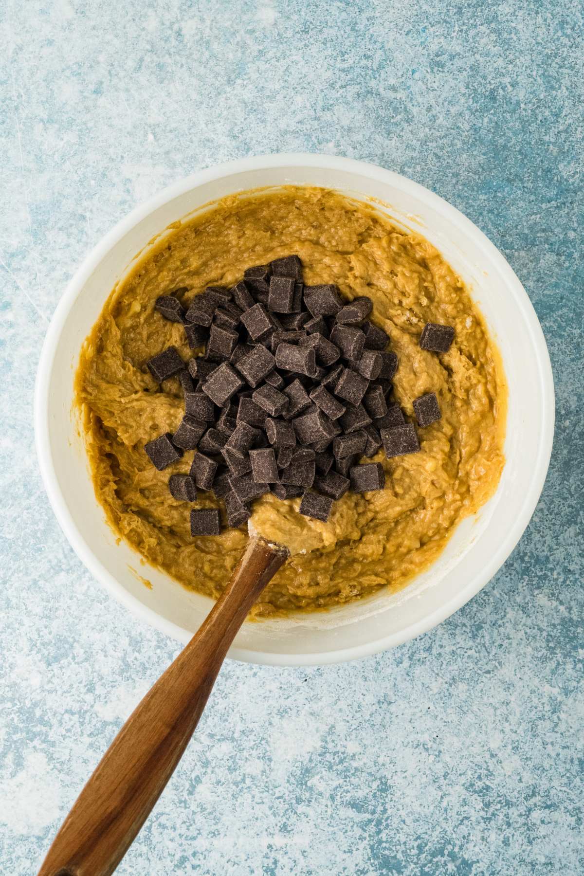 A white bowl with cookie dough and chocolate chunks being mixed with a wooden spoon on a blue background.