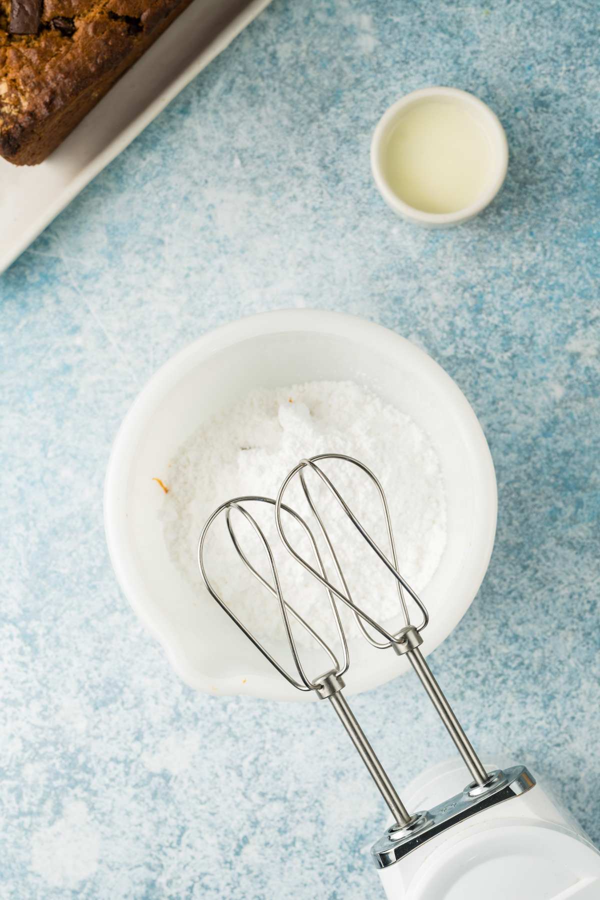 Electric hand mixer over a bowl of powdered sugar, with a small bowl of milk nearby on a blue surface.