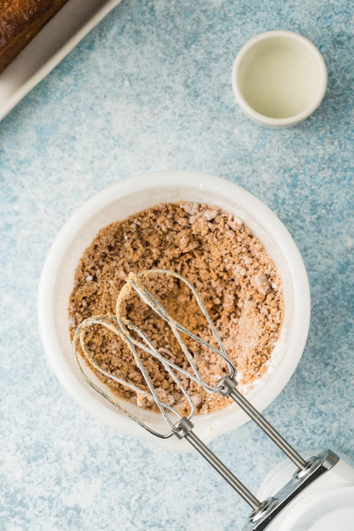 Electric mixer with beaters in a bowl of dry brown cake mix on a blue countertop; small cup nearby.