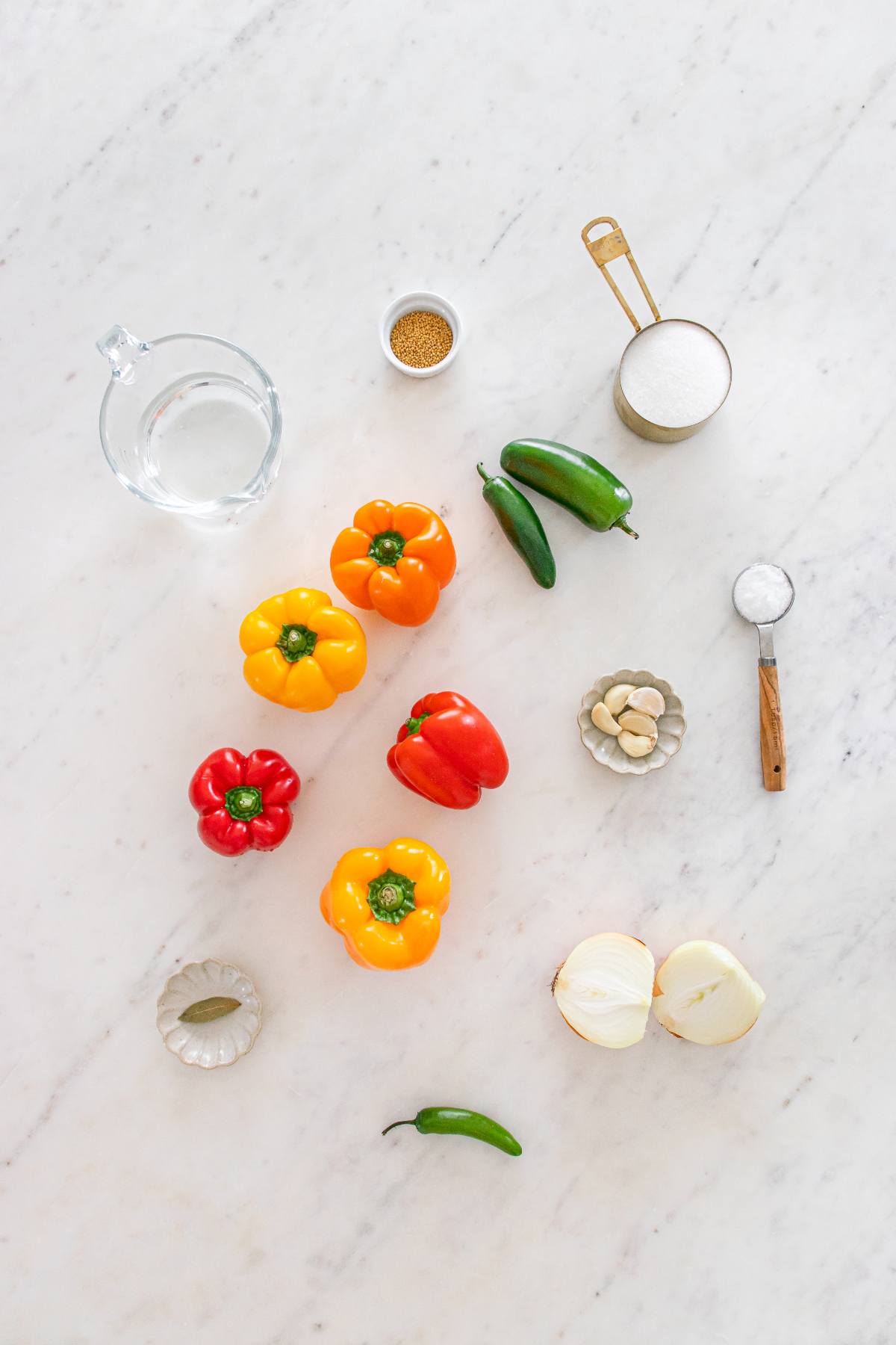 Colorful bell peppers, chilies, onion, garlic, salt, sugar, and spices arranged on a white marble surface.