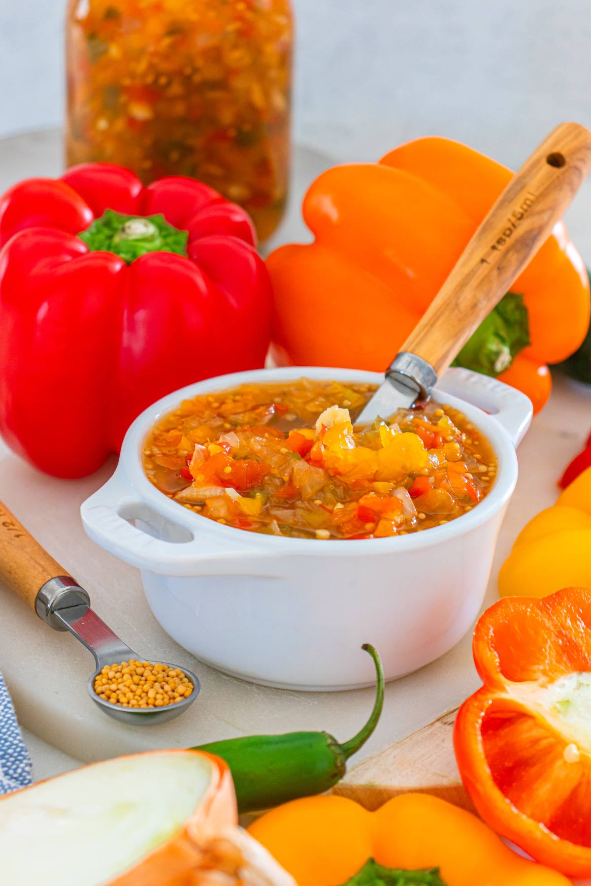 A bowl of homemade relish with chopped peppers, surrounded by fresh bell peppers and a spoon of mustard seeds.