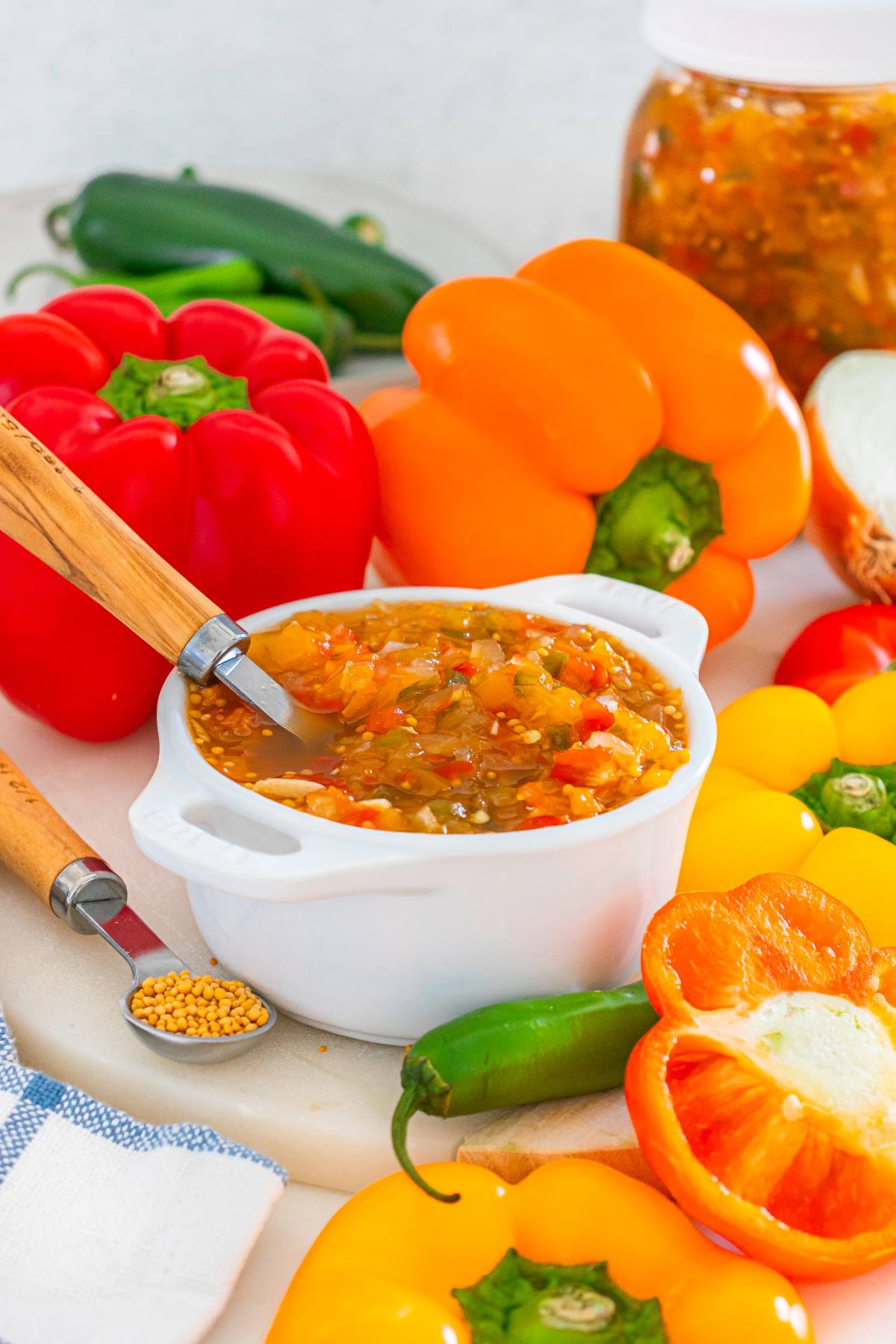 Colorful chopped pepper relish in a white bowl, surrounded by fresh peppers and vegetables on a white surface.