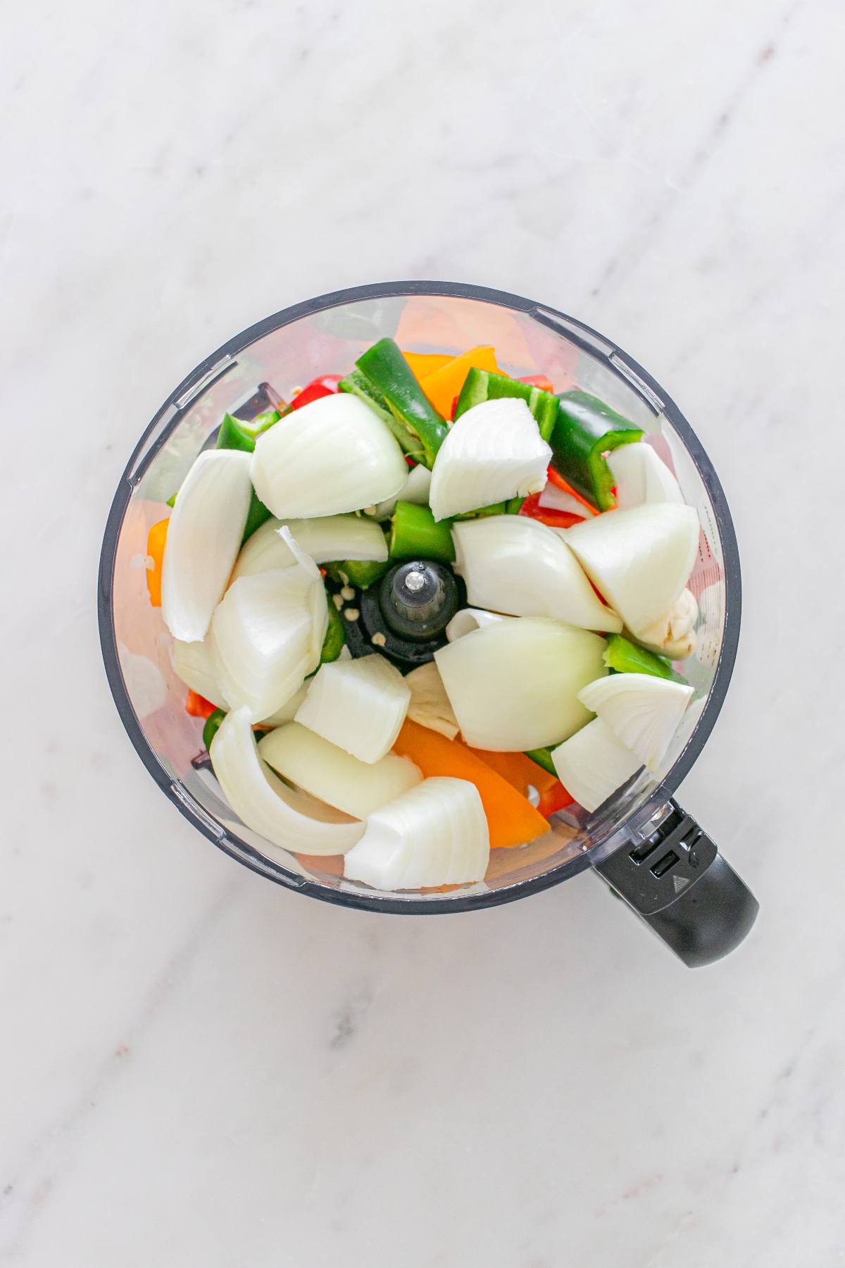 Chopped onions, peppers, and garlic in a food processor bowl on a white marble surface.