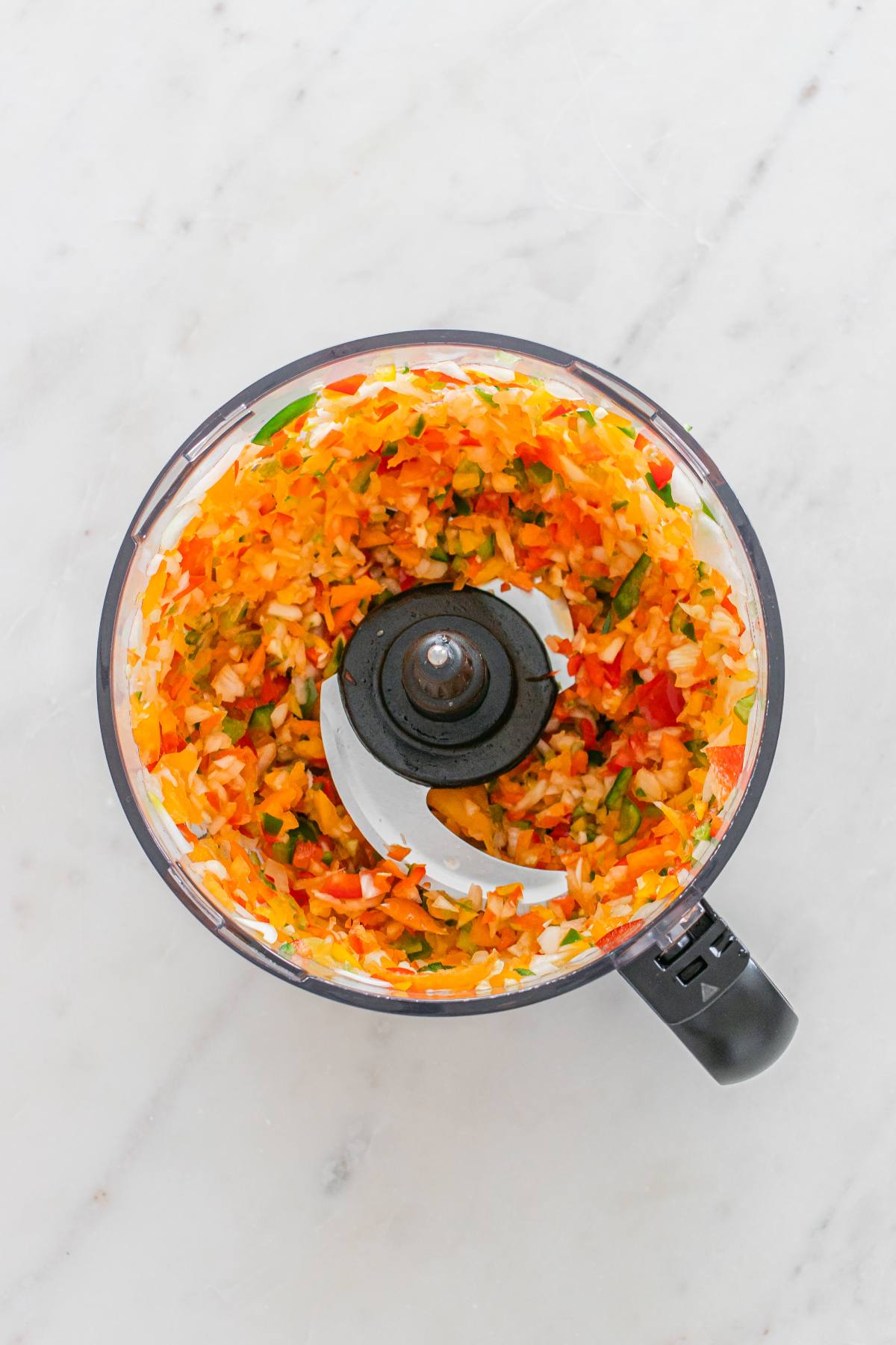 Chopped colorful vegetables in a food processor bowl on a white marble countertop.