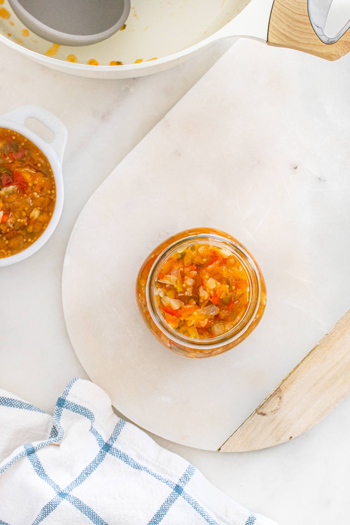 A jar of homemade relish on a white cutting board with a dish towel and pot nearby.