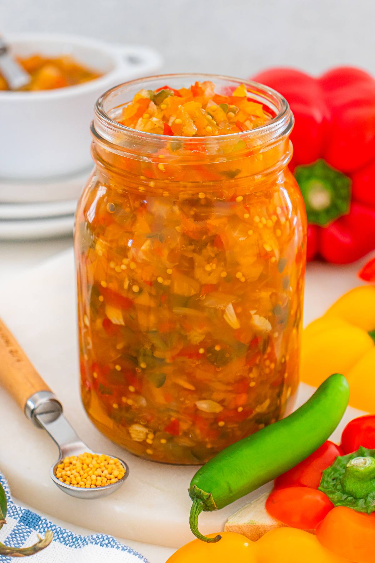 A jar of homemade relish with chopped vegetables, surrounded by fresh peppers and a green chili on a white surface.