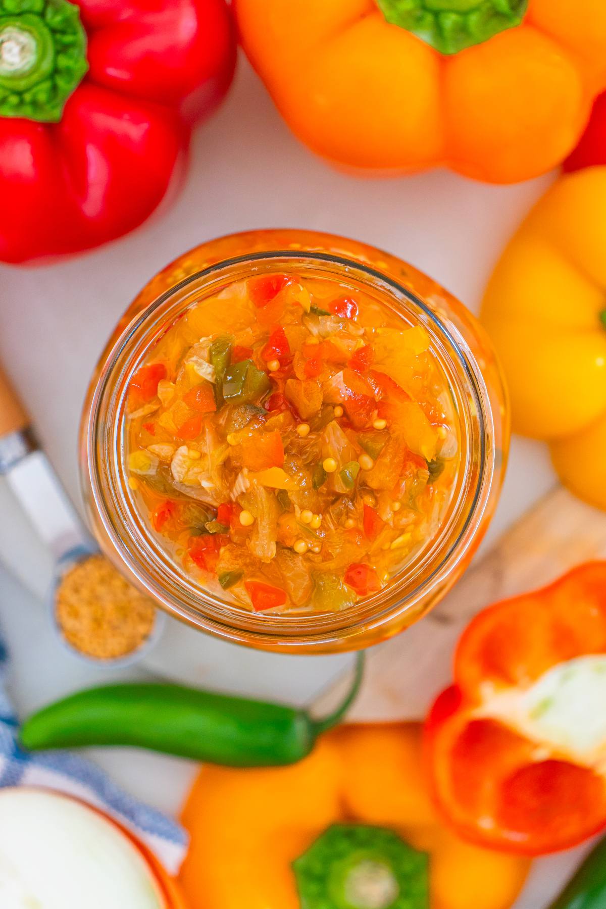 Jar of colorful homemade relish surrounded by red, orange, and yellow bell peppers, and a green chili.