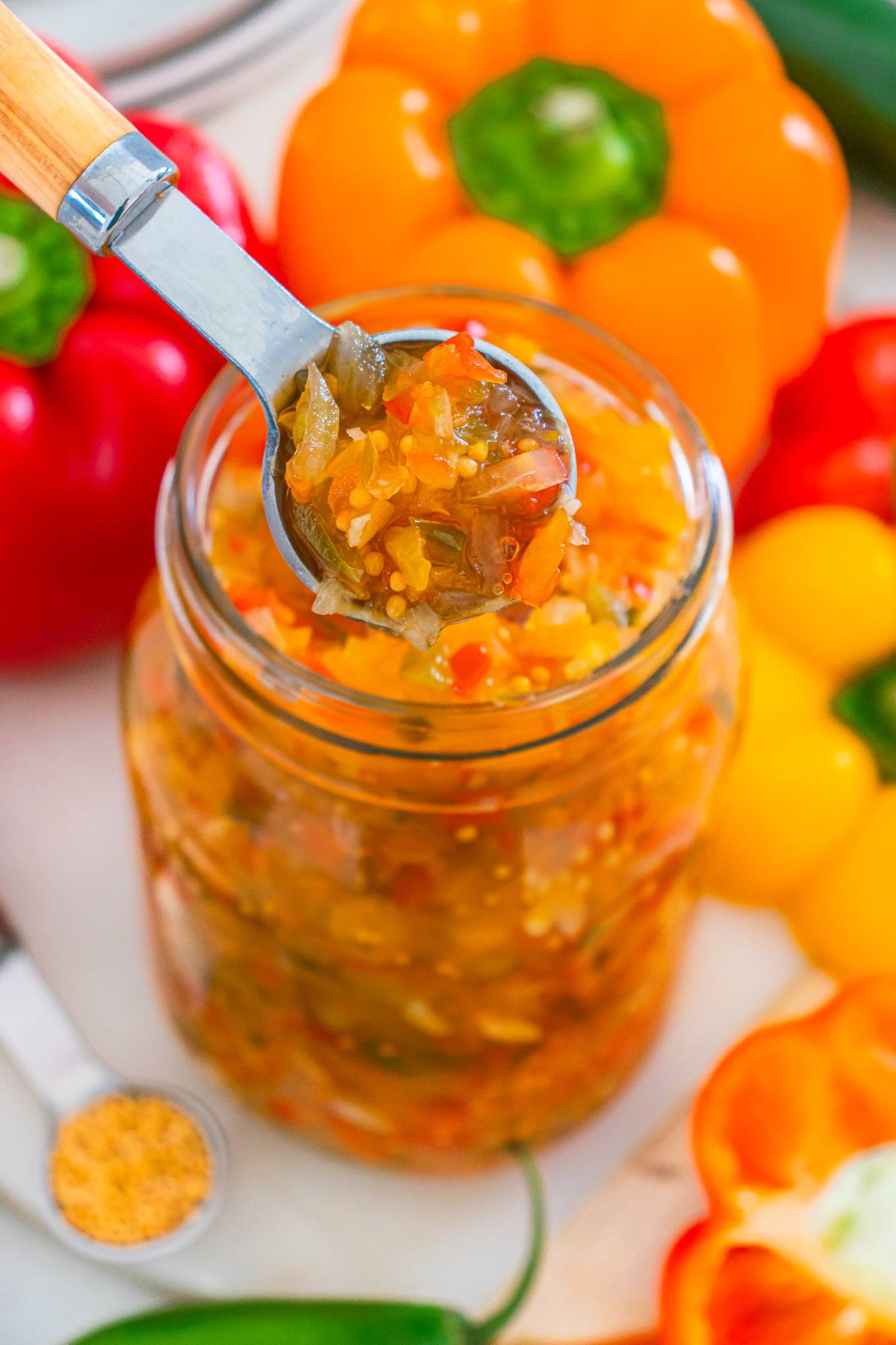 A spoon scooping homemade relish from a jar, surrounded by colorful bell peppers.