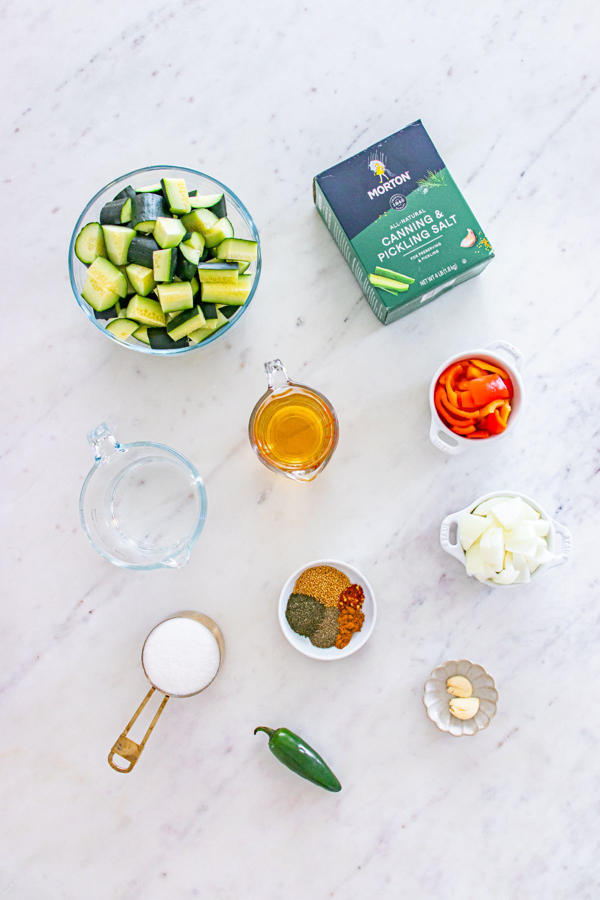 Cucumber, peppers, onion, jalapeño, garlic, spices, sugar, vinegar, and salt arranged on a marble surface.