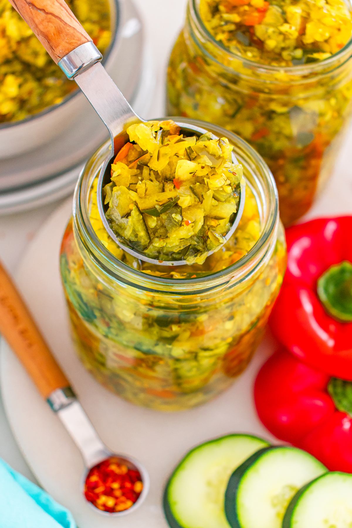 A spoon scoops relish from a jar, with cucumber, red pepper, and dried chili flakes nearby.