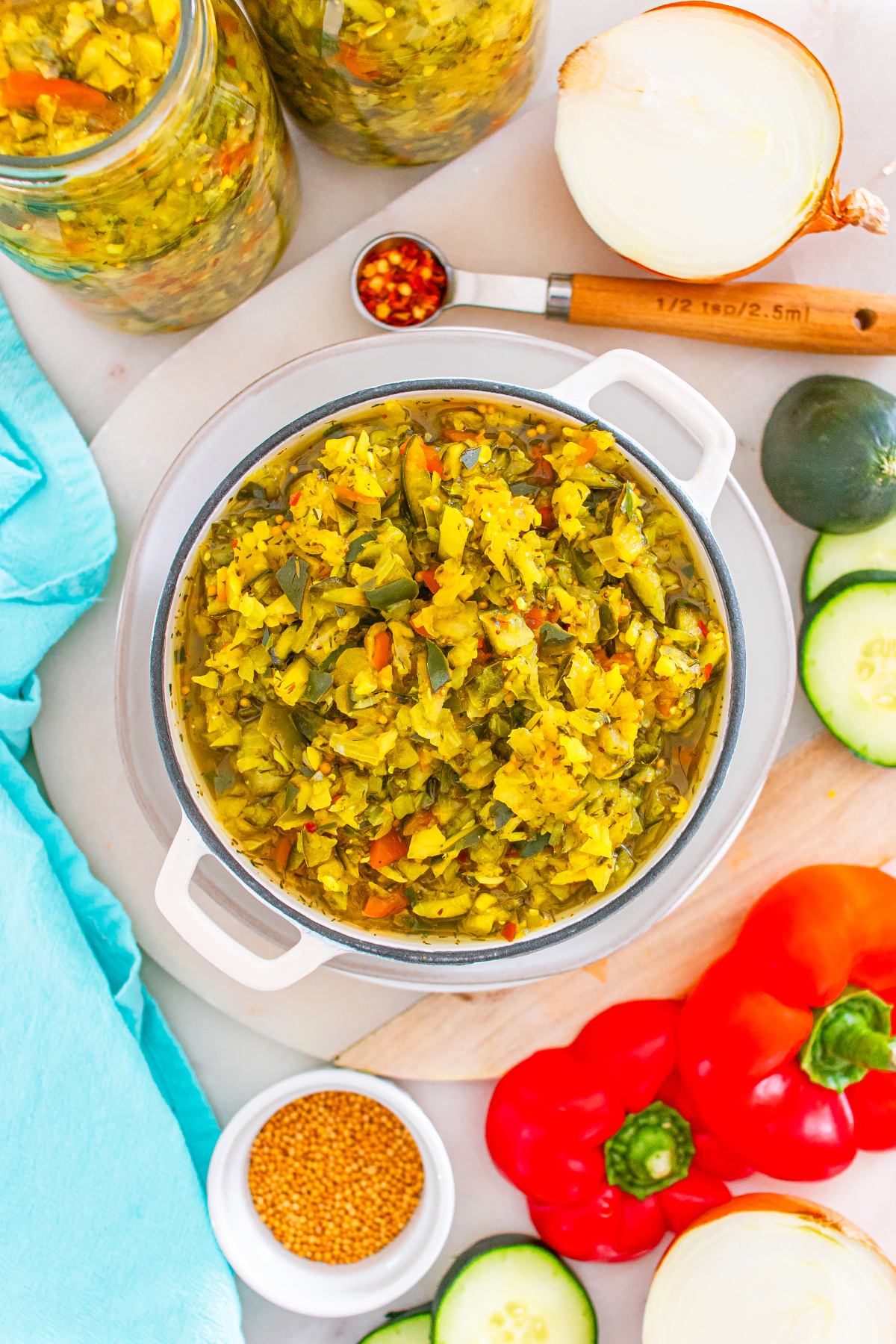 A bowl of homemade relish surrounded by jars, cucumbers, peppers, onions, and spices on a countertop.