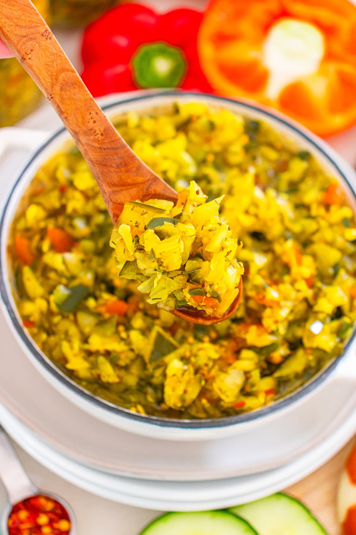 A wooden spoon lifts bright yellow relish from a white bowl, surrounded by colorful vegetables.