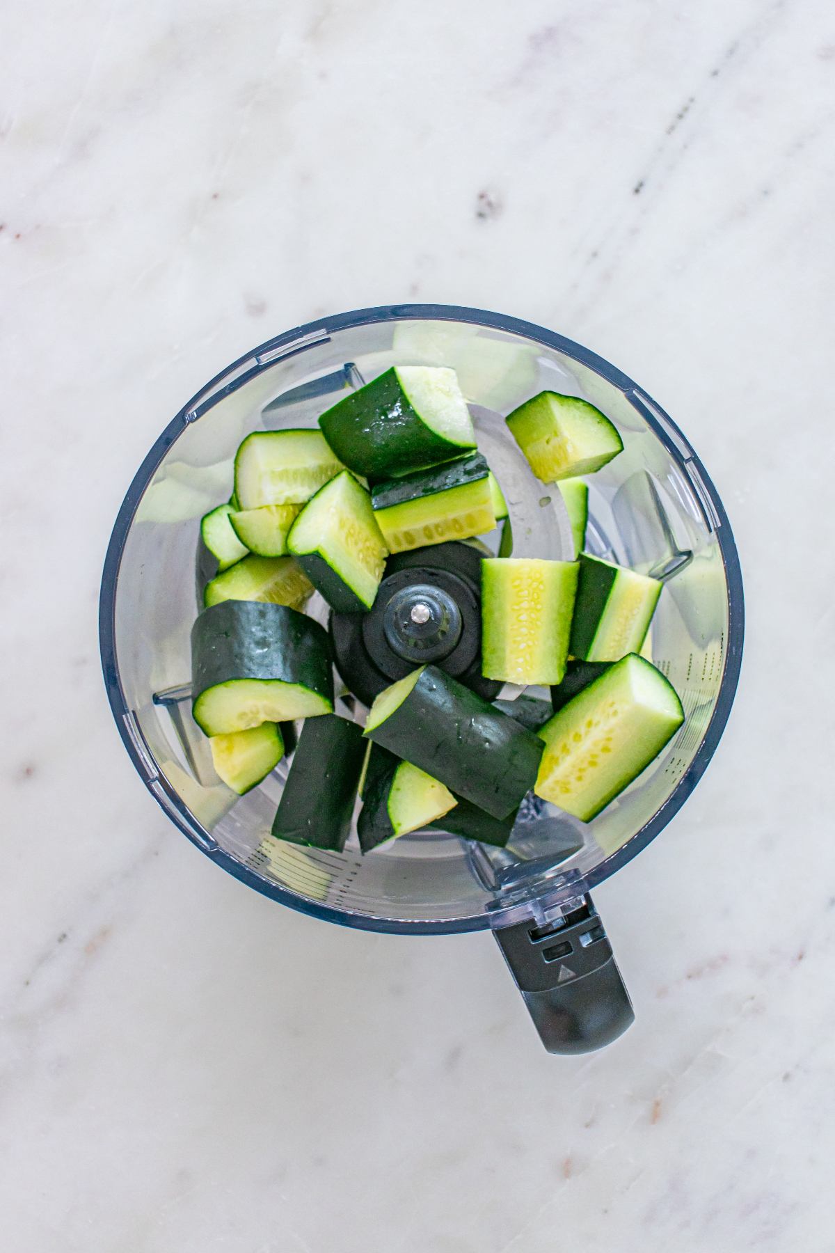 Chopped cucumber pieces in a food processor bowl on a white marble surface.