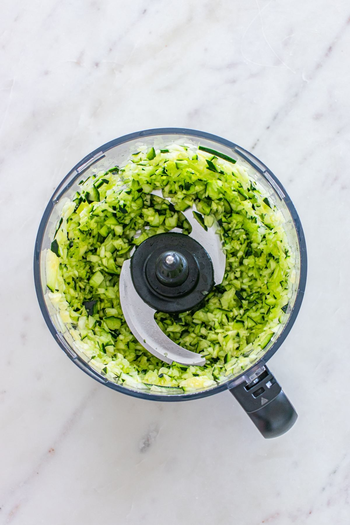 Chopped cucumber in a food processor bowl, viewed from above on a white marble surface.