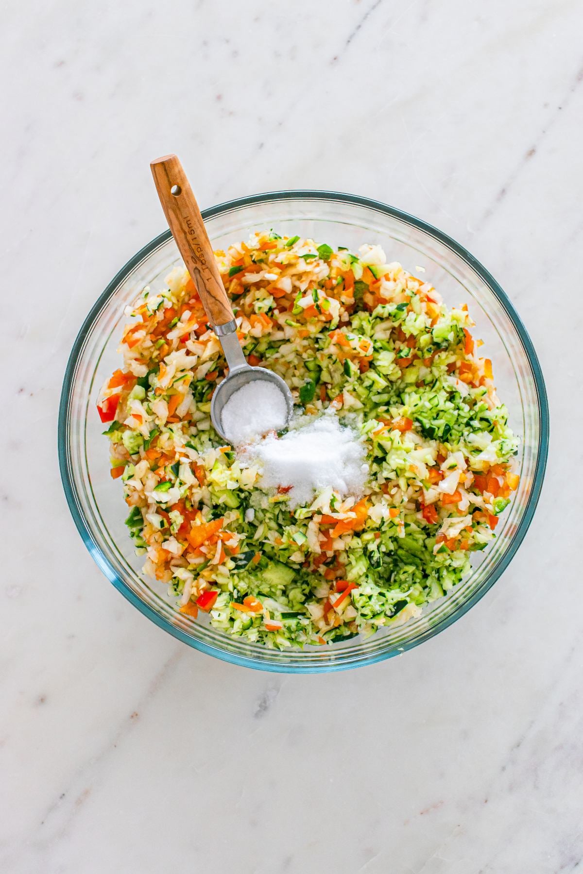 A glass bowl of chopped vegetables with a spoonful of salt on top, on a white marble surface.