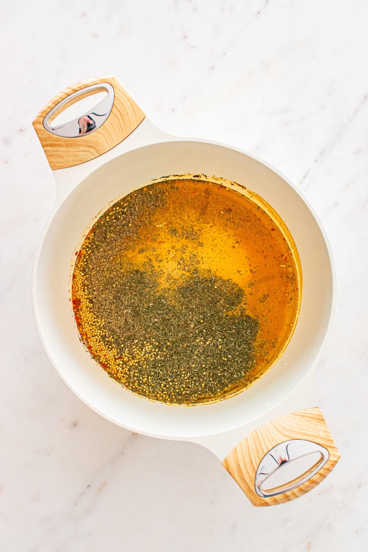 White pot with yellow broth and herbs on a white marble surface, photographed from above.