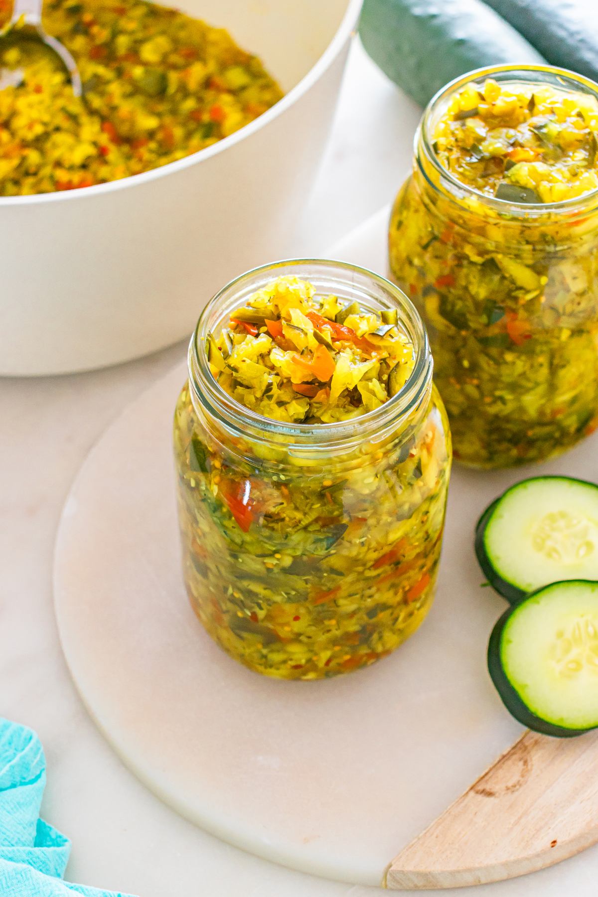 Two jars of homemade relish with chopped vegetables beside cucumber slices and a pot in the background.