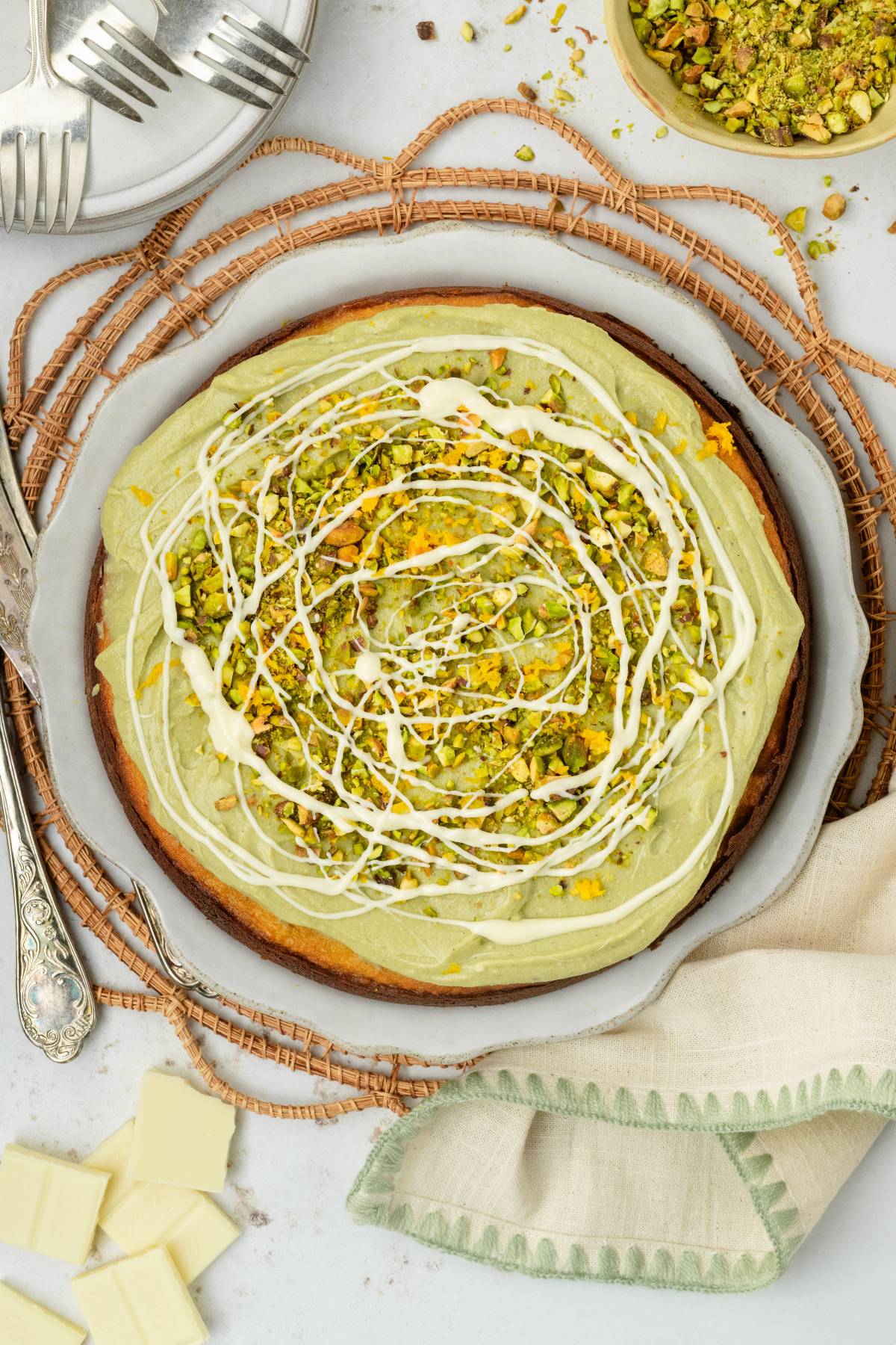Round pistachio ricotta cake with green frosting, chopped pistachios, and white chocolate drizzle on a plate, with utensils nearby.