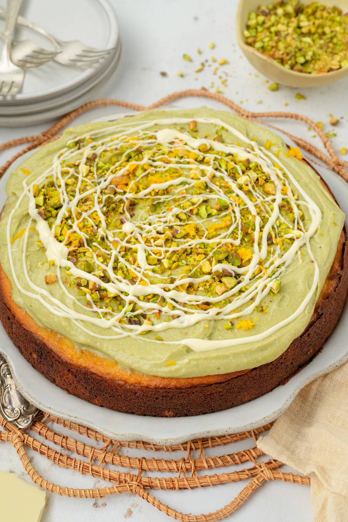A pistachio ricotta cake with green frosting, chopped pistachios, and a white icing drizzle sits elegantly on a white plate.